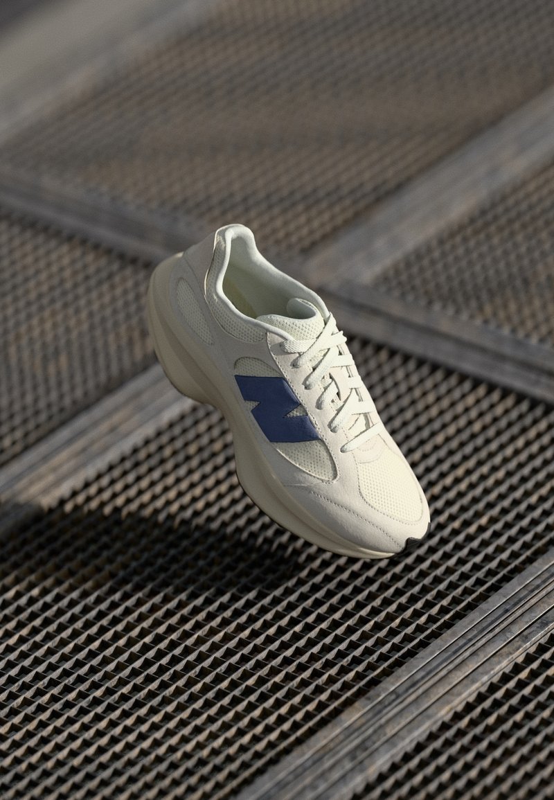 Baskets blanches avec logo latéral bleu flottant au-dessus d'une surface en grille métallique sous une lumière naturelle douce.