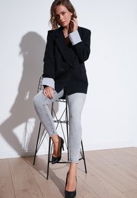 Černý dvouřadový blazer s světle modrými manžetami, kombinovaný se světle šedými skinny džíny a černými lodičkami, sedící na drátové židli.