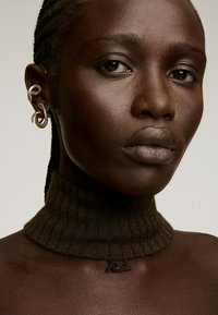 Suéter de cuello alto marrón con mangas acanaladas y un pequeño detalle del logo. El modelo lleva pendientes de aro retorcidos en oro, destacando la textura y el diseño.