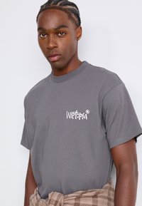 Jongeman met varkensstaartjes die een grijs "Wistopia" t-shirt en een geruite shirt om zijn middel draagt, staande tegen een witte achtergrond.