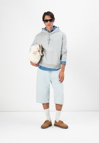 Šedá mikina s klokankou, svetlomodré denimové šortky, béžové topánky a biele ponožky, drží off-white tašku. Tmavé slnečné okuliare.
