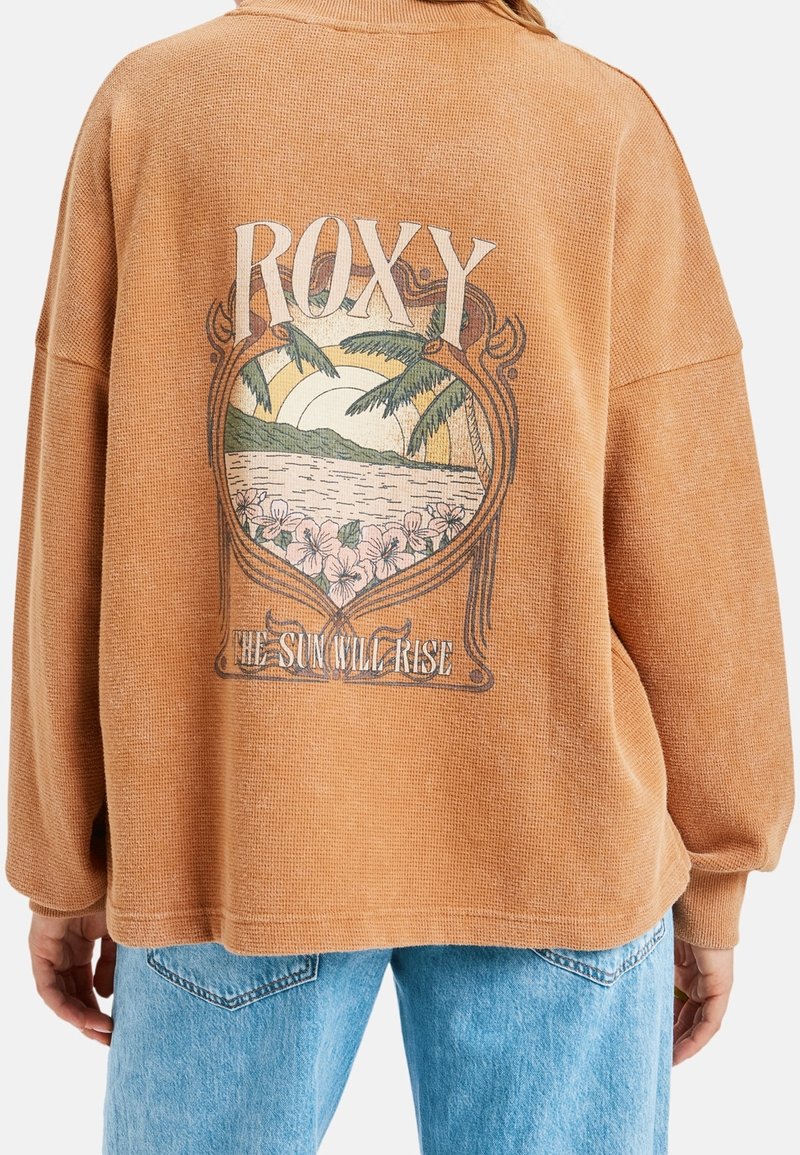 Pull marron oversize avec un imprimé tropical d’un coucher de soleil, des palmiers, des fleurs et le texte « ROXY » et « THE SUN WILL RISE » dans le dos.