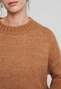 Vrouw in een getextureerde camelbruine gebreide trui en gouden creolen met schelpcharms, met haar losjes opgestoken.