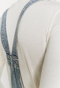 Tummat farkkuhousut säädettävillä olkaimilla, yhdistettynä vaaleanbeigeen pitkähihaiseen paitaan. Ylihaalari on klassisen mallinen ja siinä on ompeluyksityiskohtia.