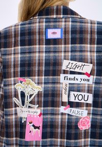 Kostkovaná košile s hnědými a námořnickými tóny, s dekoračními nášivkami, květinovým vzorem a vrstvenými textovými prvky v různých písmách.