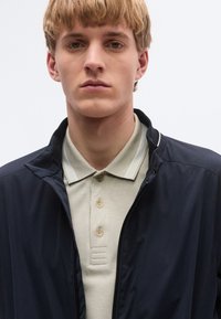 Tumesinine lukuga jakk helebeezhi poloseruudi peal, millel on tekstureeritud kangas, krae peal kolm nuppu ja peened õmblusdetailid.