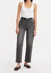 Jean taille haute gris foncé avec jambe droite, doté de cinq poches et d'une ceinture noire. Associé à un débardeur blanc et des ballerines noires.