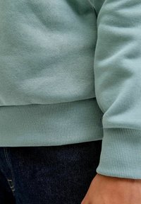 Primer plano de una persona que lleva un suéter verde claro con puños acanalados y jeans de mezclilla azul oscuro, mostrando parte del brazo y la cintura.