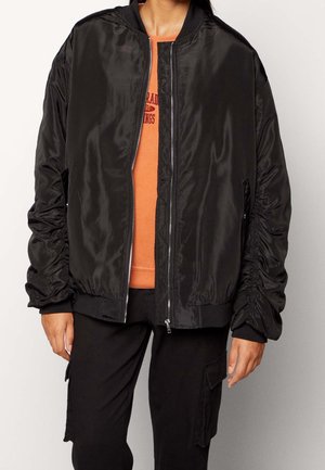 Osoba ubrana w błyszczącą, czarną kurtkę bomber z marszczonymi rękawami, na pomarańczowej bluzie i czarnych spodniach typu cargo, stojąca na jednolitym tle.