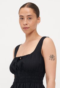 Μαύρο αμάνικο φόρεμα με τετράγωνη λαιμόκοψη, μαζεμένο μπροστινό μέρος και λάστιχο στη μέση. Περιλαμβάνει λεπτομέρεια με φιόγκο στο στήθος και λεία υφή υφάσματος.