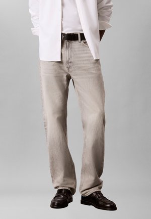 Mężczyzna w jasno beżowych, luźnych dżinsach, czarnym pasku, białej koszuli i ciemnobrązowych skórzanych butach na szarym tle.