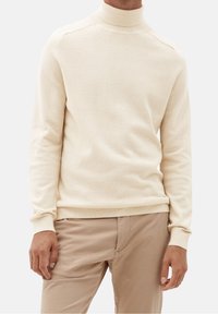 Pull à col roulé couleur crème avec un motif tricoté texturé, poignets et ourlet côtelés. Associé à un pantalon beige clair.