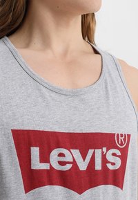 Šedý bavlněný top na ramínka s velkým červeným logem Levi's a bílým textem. Kulatý výstřih a volný střih. Měkká textura a minimální švy.