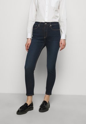 Person trägt dunkle Skinny-Jeans, weißes langärmeliges Hemd mit Knöpfen und schwarze Tassel-Loafer, steht vor einem schlichten Hintergrund.