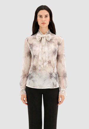 Donna che indossa una camicetta trasparente a fiori con maniche lunghe e un grande fiocco annodato al collo, abbinata a pantaloni neri a gamba dritta.