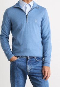 Pull bleu clair à fermeture éclair en tissu doux avec manches longues et poignets côtelés, associé à un jean bleu décontracté. Présente un écusson logo sur la poitrine.