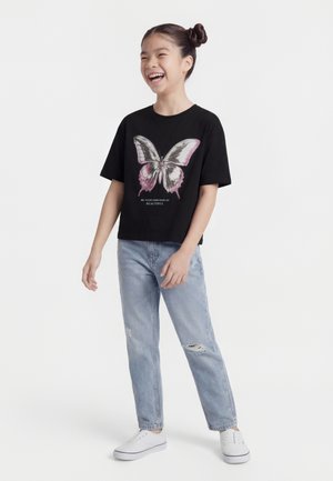 Fille riant, portant un t-shirt noir avec un grand papillon et du texte, un jean clair déchiré, et des baskets blanches, debout devant un fond uni.