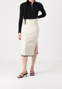 Frau mit einem gerippten cremefarbenen Midiskirt mit seitlichem Schlitz, einem schwarzen Langarmoberteil mit Reißverschlusskragen und schwarzen Strappy-High-Heels.
