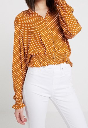 Frau trägt eine blusenartige Bluse in Senfgelb mit weißen Punkten und gekräuseltem Saum, kombiniert mit hoch taillierten weißen Hosen.