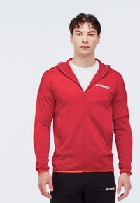 Red zip-up athletic jacket with hood, long sleeves, lightweight material, and contrasting orange accents. Logo on the left chest area.