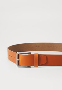 Ceinture en cuir d'un orange vif avec une boucle en métal argenté. Elle présente une texture lisse et plusieurs trous d'ajustement. Le logo de la marque est gravé et visible.