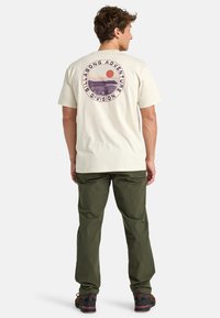 Camiseta beige clara con un gráfico circular que muestra un atardecer y el océano, combinada con pantalones verdes y calzado oscuro.