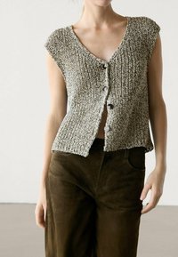 Gilet lavorato a maglia in tonalità grigio-marrone, con scollatura a V, maniche corte e tre bottoni sul davanti. Abbinato a pantaloni marrone scuro.