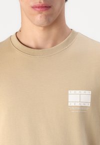 Beige cotton t-shirt with a crew neck, featuring a small white logo on the left chest. Smooth texture with a simple design.