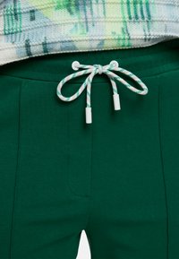 Groene broek met trekkoord, gestrikt met een wit-groen patroonkoord, gecombineerd met een meerkleurige gebreide trui.