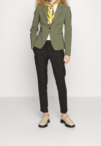 Blazer su misura verde oliva, pantaloni neri slim-fit e scarpe beige stringate. Top bianco sotto, accento di sciarpa gialla. Tessuto liscio.