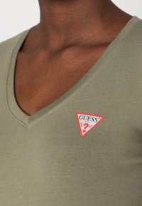 Olive green, fitted V-neck t-shirt made of smooth fabric, featuring a red and white triangular "GUESS" logo patch on the left chest.