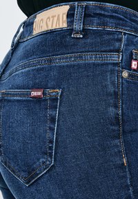 Prim-plan al buzunarului din spate al unei perechi de blugi din denim albastru închis, cu etichetă de piele Big Star de culoare maro deschis și etichetă de material roșu cu logo stea.