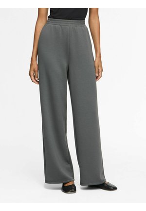 Personne portant un pantalon large gris charbon avec une taille élastique et des chaussures plates noires, debout devant un fond blanc.