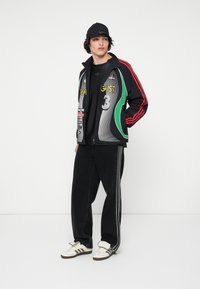 Ung mann som har på seg svart cap, fargerik Adidas treningsjakke, svart t-skjorte, svarte bukser med sidestriper, og hvite Adidas sneakers, står på siden.