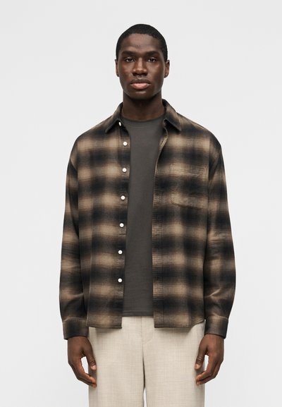 Homme portant une chemise à carreaux marron et noire boutonnée par-dessus un t-shirt gris foncé et un pantalon beige clair, debout devant un fond blanc uni.