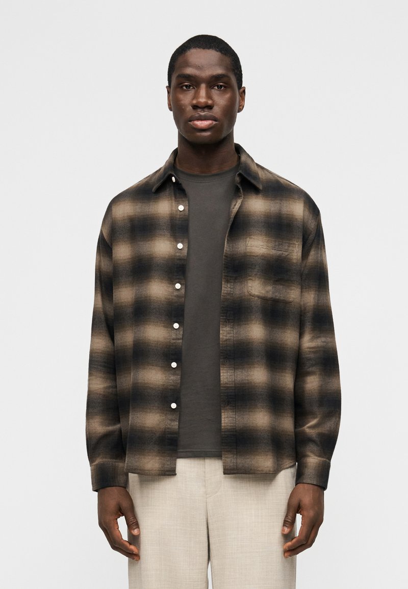 Άνδρας που φοράει καρό πουκάμισο σ ε καφέ και μαύρο πάνω από μαύρο γκρι μπλουζάκι και ανοιχτό μπεζ παντελόνι, στέκεται μπροστά σε απλό λευκό φόντο.