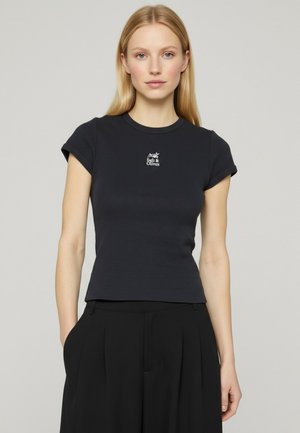 Młoda kobieta o blond włosach nosi czarną obcisłą bluzkę z krótkim rękawem z małym białym logo oraz luźne czarne spodnie z wysokim stanem, stoi na tle jednolitego tła.