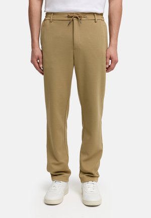 Pantalones beige de hombre con cordón y bolsillos laterales, combinados con zapatillas blancas y una camisa blanca, mostrados desde la cintura hasta los pies sobre un fondo blanco.