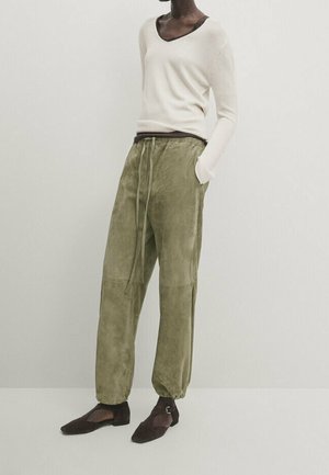 Personne portant un haut beige à manches longues, un pantalon vert olive avec cordon de serrage et ourlets élastiques, ainsi que des chaussures plates marron foncé avec des brides à la cheville, debout contre un mur uni.
