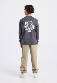 Harmaa huppari, jossa on valkoinen graafinen kuvio selässä, yhdistettynä beigeihin joogapantoihin ja valkoisiin lenkkareihin. Asu on rento ja siinä on elastiset hihansuut.