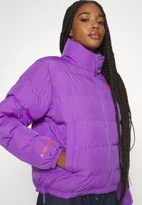 Violetinė pūkinė striukė su aukštu apykakle, elastingomis rankogaliais ir priekiniais užtrauktukais. Ant rankovės išsiuvinėtas Helly Hansen logotipas oranžine spalva.