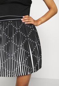 Zwarte geplooide rok met witte verticale strepen en herhaaldelijk "GUESS" logo. De tailleband heeft een contrasterende witte biezing. Gladde textuur.