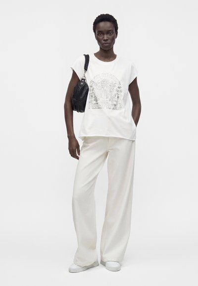 Femme en haut sans manches blanc à motif graphique et pantalon large, tenant un sac à épaule noir, debout devant un fond blanc uni.