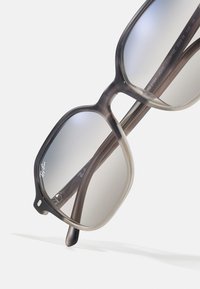 Óculos de sol com uma armação cinza translúcida, de formato quadrado, e lentes degradê. As hastes apresentam uma marcação sutil e uma textura suave.