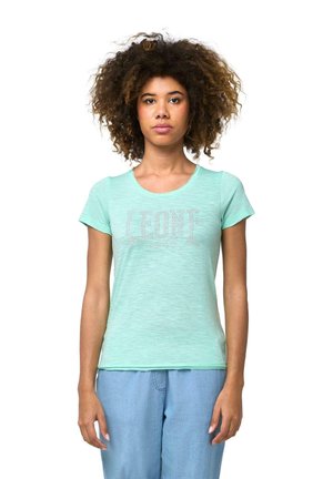 Une femme aux cheveux bouclés et volumineux se tient face à l’objectif, vêtue d’un tee-shirt vert menthe à motif et d’un pantalon bleu clair, sur fond blanc.