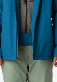 Uomo che indossa una giacca blu sopra un maglione grigio con zip e pantaloni verde chiaro, mani rilassate ai lati, giacca aperta.