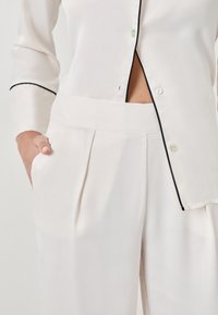 Pantalons plissés blancs taille haute avec une main dans la poche et une chemise blanche partiellement boutonnée avec des bordures noires sur les poignets et la couture avant.