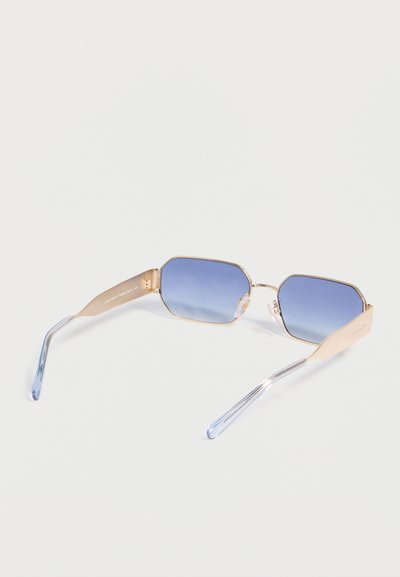 Γυαλιά ηλίου με χρυσή κορνίζα, εξαγωνικούς φακούς με μπλε διαβαθμισμένο χρώμα και διαφανείς μπλε άκρες στους βραχίονες πάνω σε λευκό φόντο.