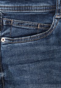 Tummansinisiä farkkuja, joissa on etutasku, näkyvät ompeleet kontrastivärisessä kultaisessa sävyssä ja metallinen niitti.