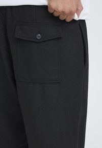 Svarte bukser laget av teksturert stoff med en sidelomme med klaff og knappelukking. Avslappet passform med en glatt finish.